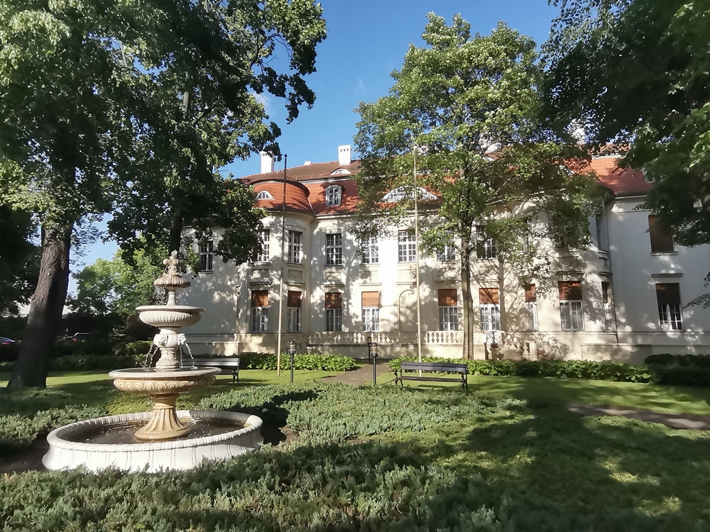 Zdjęcie przedstawia park, kamienną fontannę i w tle widoczny budynek pałacowy. Jest pałac Alfreda Biedermanna 