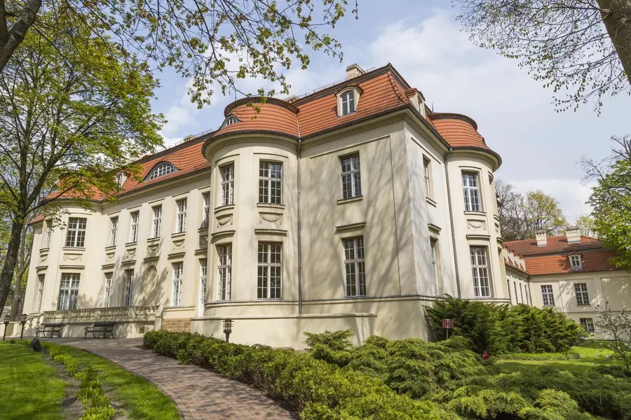 Na zdjęciu widoczny jest duży budynek, który wygląda jak pałac. Obiekt stoi wśród drzew. Widać, że jest słoneczny dzień.