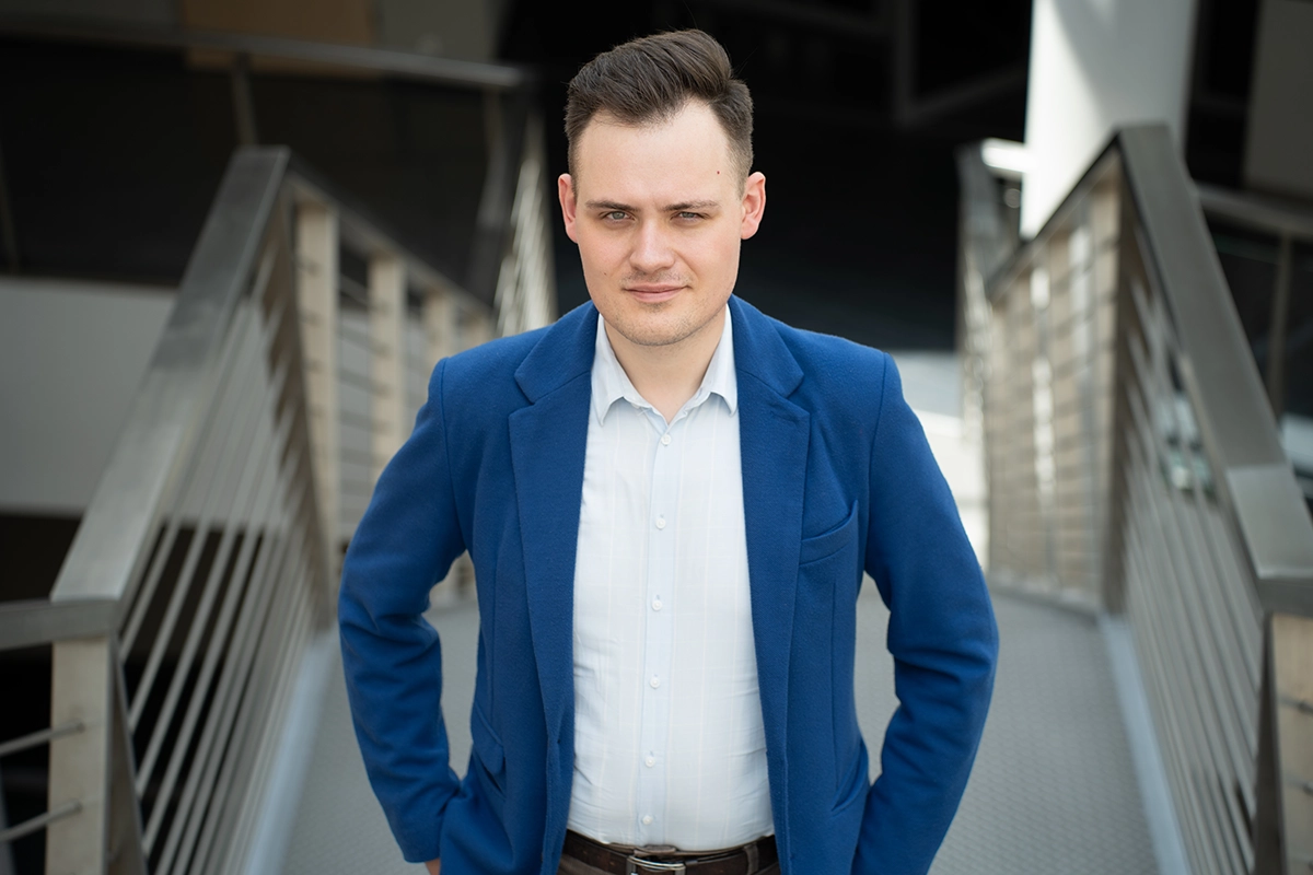 Młody mężczyzna w niebieskiej marynarce i koszuli na tle schodów.