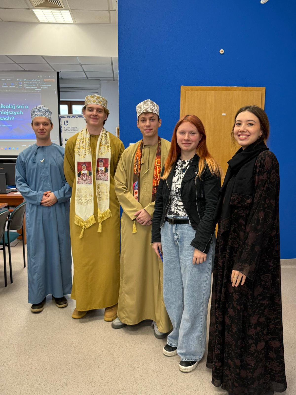 Pięć osób stoi w pomieszczeniu przed niebieską ścianą i drewnianymi drzwiami. Trzy osoby mają na sobie tradycyjne stroje bliskowschodnie, w tym długie szaty i wzorzyste nakrycia głowy. Dwie pozostałe są ubrane w nowoczesne, codzienne ubrania. W tle widoczny jest ekran z prezentacją w języku polskim.