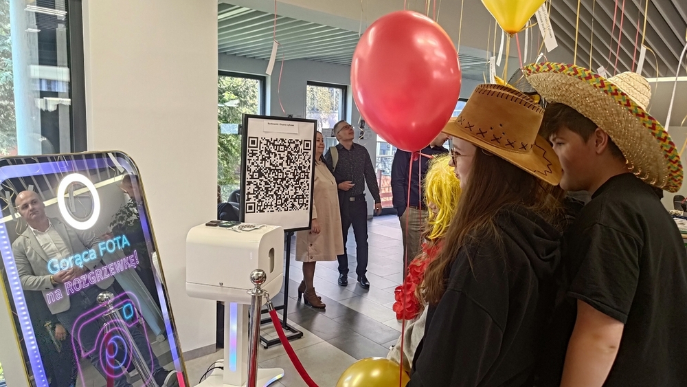 drzwi otwarte 2025 - dwie dziewczyny i chłopak trzymają balony z helem i pozują w słomianych kapeluszach do fotobudki w kształcie dużego telefonu komórkowego z napisem "gorąca fota na rozgrzewkę"
