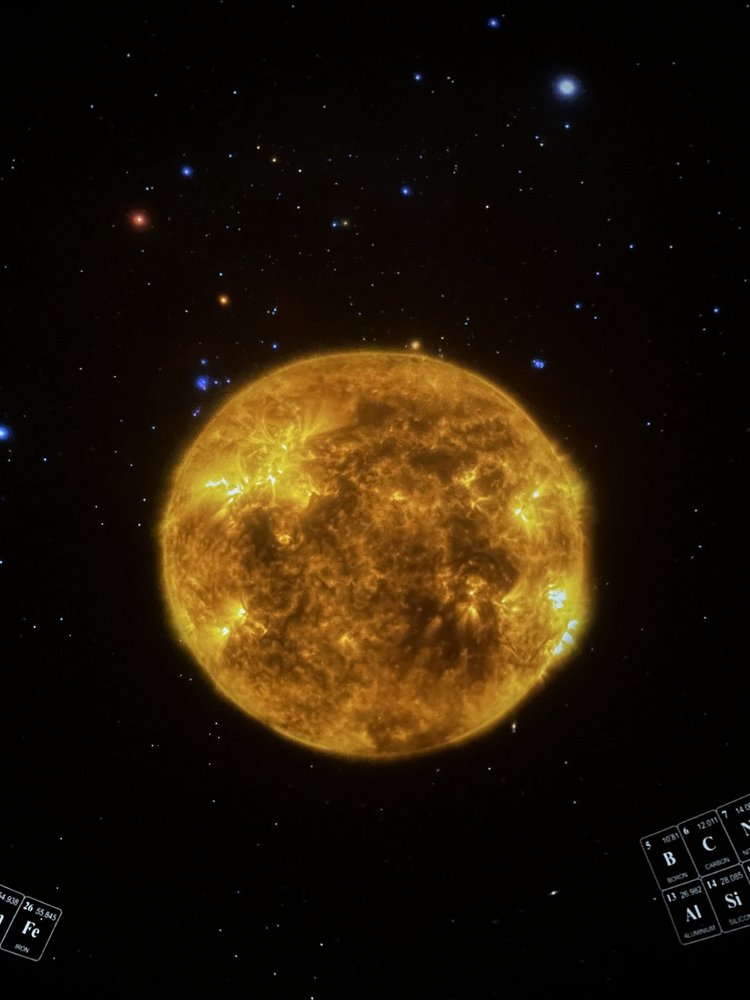 Na środku obrazu znajduje się duża, jasnożółta kula przedstawiająca Słońce. Jego powierzchnia jest nierówna i dynamiczna – widoczne są jaśniejsze i ciemniejsze obszary, przypominające gorące, wijące się struktury plazmy. W kilku miejscach pojawiają się wyraźne, intensywnie świecące punkty, które wyglądają jak rozbłyski słoneczne. Tło zdjęcia jest czarne i wypełnione licznymi gwiazdami w różnych kolorach: białymi, niebieskimi, pomarańczowymi i czerwonymi. W dolnych rogach kadru widać fragmenty schematycznych tablic – przypominają elementy tablicy okresowej pierwiastków. Całość przypomina naukową wizualizację lub kadr z filmu edukacyjnego o Słońcu i strukturze Wszechświata.