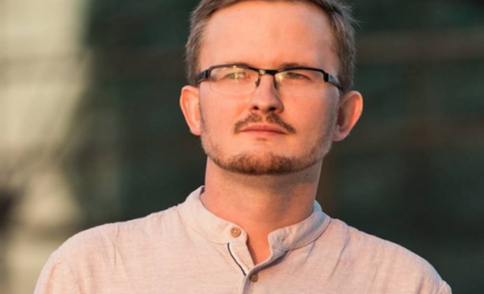 mężczyzna w okularach i jasnej koszuli na tle budynku