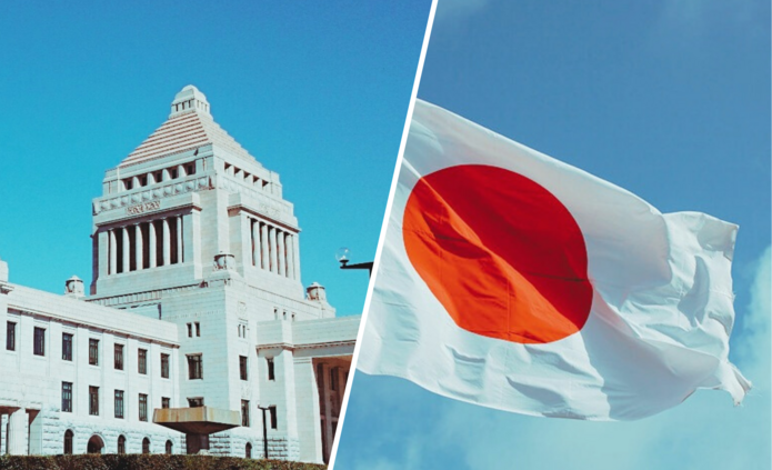 Po lewej stronie biały budynek na tle nieba, po prawej stronie powiewająca flaga Japonii na tle nieba