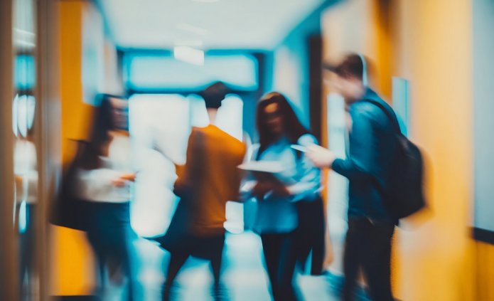 Grupa kilku osób w ruchu w korytarzu; jedna osoba trzyma dokumenty, inna zagląda w telefon. Wnętrze jest oświetlone światłem dziennym i sztucznym, występują ciepłe i chłodne barwy. Postacie są nieostre, scena sugeruje swobodną, codzienną sytuację.