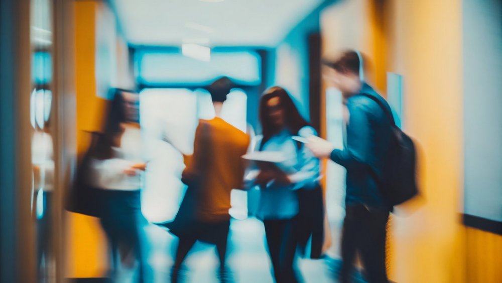 Grupa kilku osób w ruchu w korytarzu; jedna osoba trzyma dokumenty, inna zagląda w telefon. Wnętrze jest oświetlone światłem dziennym i sztucznym, występują ciepłe i chłodne barwy. Postacie są nieostre, scena sugeruje swobodną, codzienną sytuację.