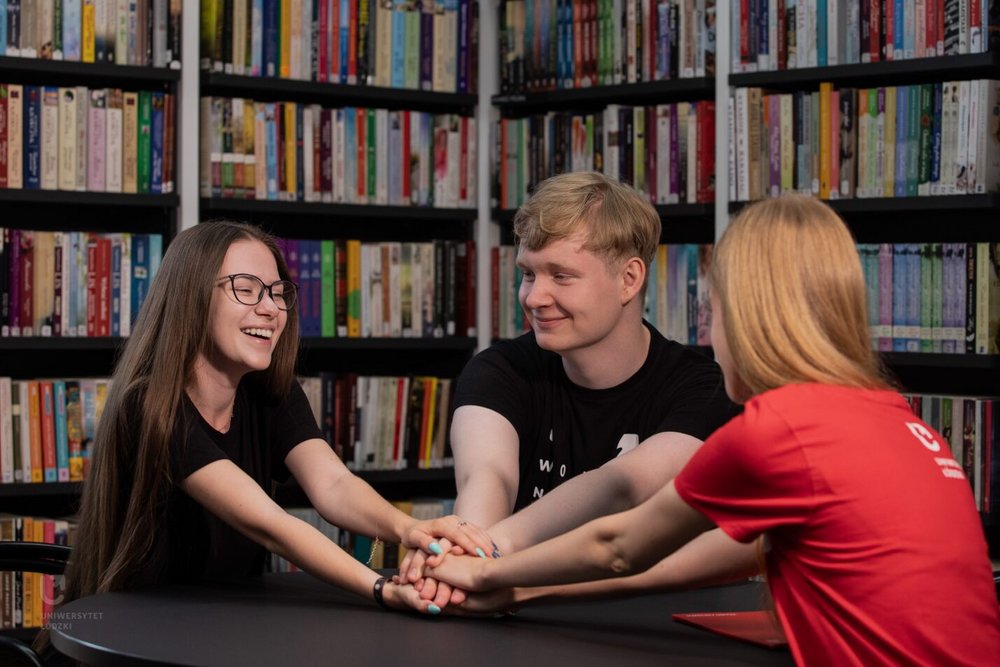 Kolorowe zdjęcie przedstawiające troje uśmiechniętych studentów siedzących na tle bibliotecznego księgozbioru. Na zdjęciu znajdują się dwie kobiety i jeden mężczyzna. Cała trójka trzyma się za ręce w znaku gotowości do wspólnego podjęcia wyzwania. Zdjęcie ma ciepły i jednocześnie motywacyjny charakter