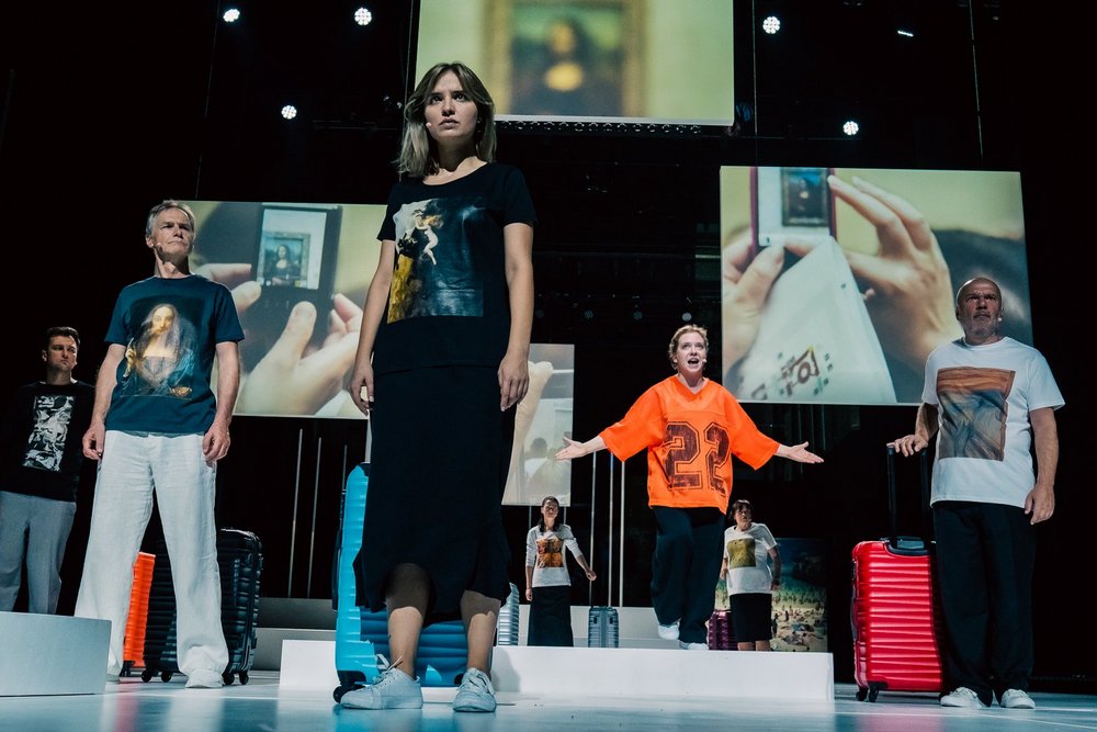 Na zdjęciu widoczna jest grupa osób stojących na scenie. Osoby są ubrane jak turyści, mają walizki. Za nimi wiszą różnej wielkości plansze na których są zdjęcia z różnych turystycznych miejsc.