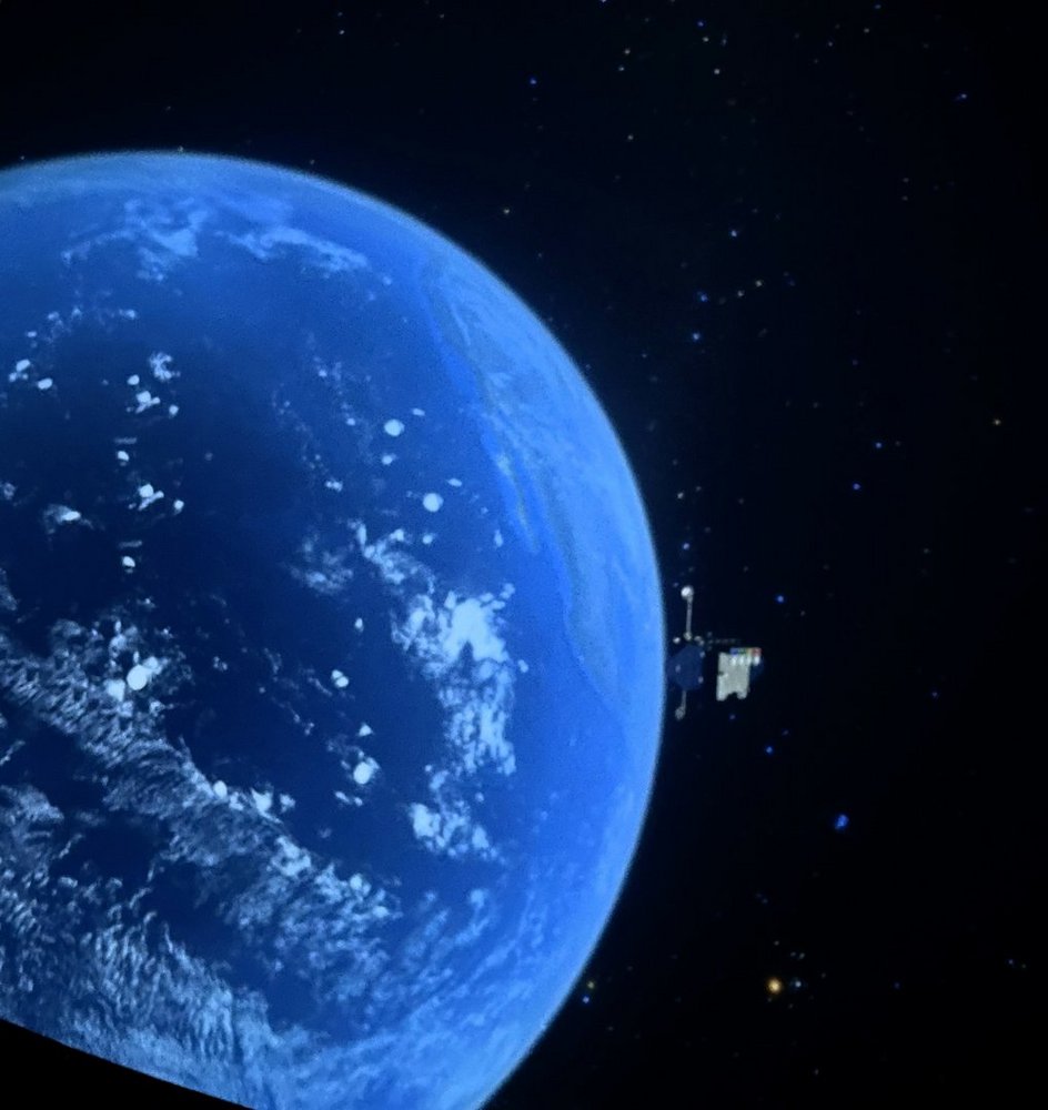 Zdjęcie przedstawia fragment Ziemi widzianej z przestrzeni kosmicznej. Widzimy dużą, zakrzywioną powierzchnię planety, w większości pokrytą oceanem o intensywnie niebieskim kolorze. Na powierzchni widać białe chmury, rozproszone w różnych miejscach nad wodą. Tło jest czarne, pełne drobnych, jasnych punktów przypominających gwiazdy. Po prawej stronie zdjęcia, na tle gwiazd, znajduje się niewielki obiekt – wygląda jak sztuczny satelita z panelami słonecznymi lub antenami wystającymi z jego boku. Całość sprawia wrażenie spokojnej sceny obserwowanej z orbity okołoziemskiej.