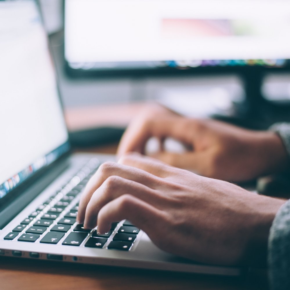 Na pierwszym planie znajduje się laptop, na którego klawiaturze znajdują się palce.