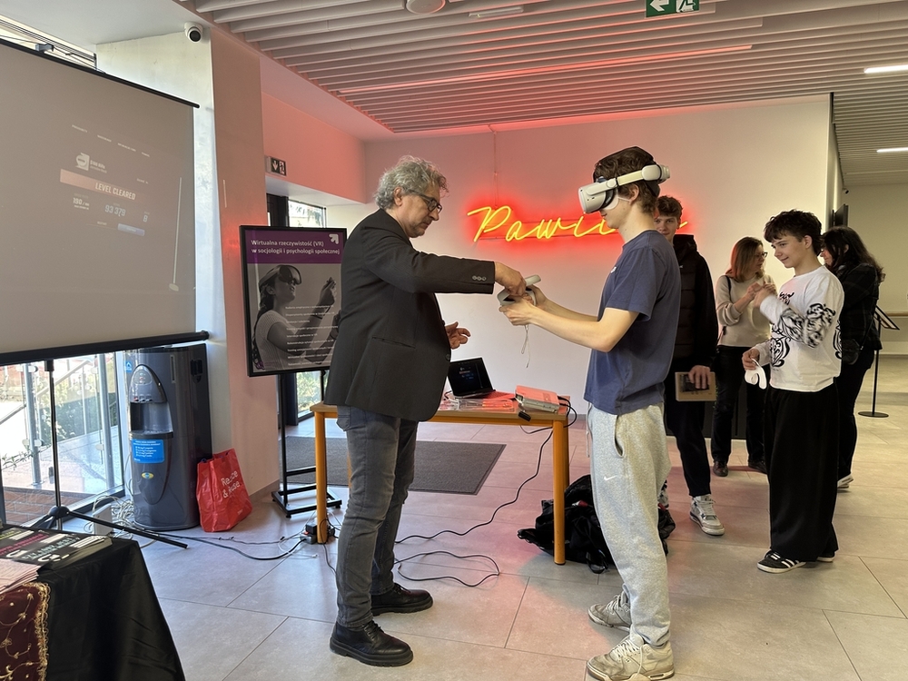 drzwi otwarte 2025 - wykładowca stoi przed stoiskiem socjologii i pokazuje uczniowi jak uruchomić google VR, które uczeń testuje na sobie