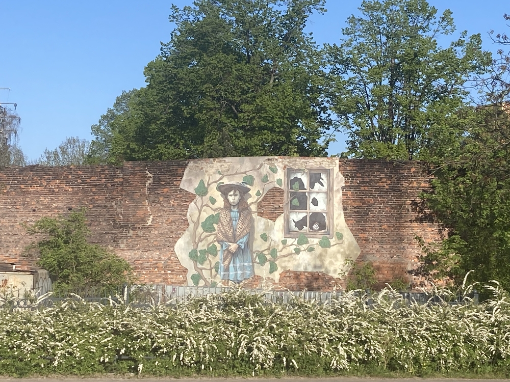 Mural przedstawiający młodą kobietę namalowany na ceglanym murze, stojącym wśród zieleni