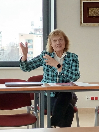 rozległa przestrzeń auli, kobieta siedzi, wygłasza wykład, gestykuluje