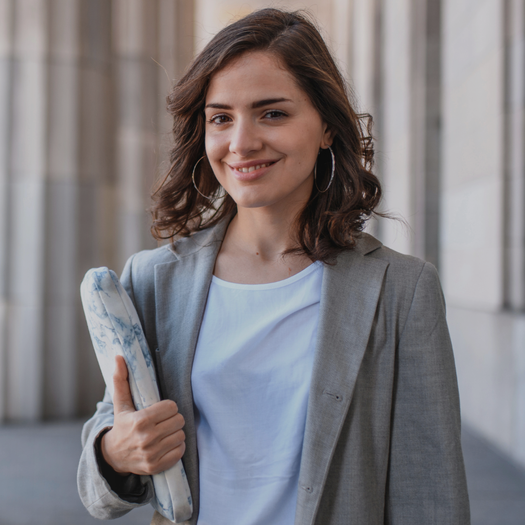 Młoda kobieta o brązowych, falowanych włosach i delikatnym uśmiechu stoi na tle monumentalnych, kamiennych kolumn. Ma na sobie szarą marynarkę i białą bluzkę, w ręku trzyma zwinięty materiał lub etui w marmurkowy wzór, pokazując kciuk uniesiony w geście aprobaty. Nosi duże, okrągłe kolczyki i sprawia wrażenie pewnej siebie oraz profesjonalnej.